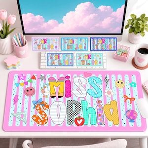 May include: A rectangular desk mat featuring a pink and light blue striped background with the text MISS SOPHIA in large, multi-patterned letters. The design includes various icons like a duck, apple, globe, smiley faces, and geometric shapes.