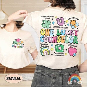 Puede incluir: Una camiseta blanca con un dise&ntilde;o gr&aacute;fico colorido que dice "One Lucky Counselor". El dise&ntilde;o incluye arco&iacute;ris, corazones, estrellas y tr&eacute;boles. El texto "Miss Sophia" est&aacute; en la parte delantera de la camiseta.