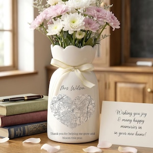 May include: A white ceramic vase with a heart-shaped map design, filled with pink and white flowers. The vase has a ribbon tied around it and the text "Mrs. Wilson" and "Thank you for helping me grow and bloom this year." A card reads "Wishing you joy & many happy memories in your new home!"