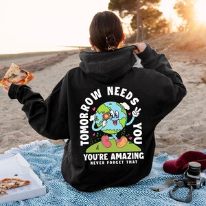 May include: Black hoodie with a graphic of a smiling cartoon Earth holding a flower and peace sign. The text "Tomorrow Needs You" is written around the Earth and "You're Amazing Never Forget That" is written below the Earth.