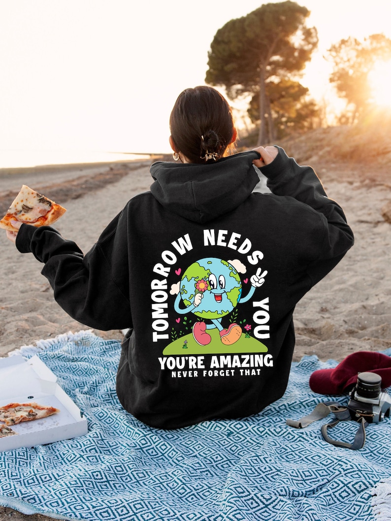 May include: Black hoodie with a graphic of a smiling cartoon Earth holding a flower and peace sign. The text "Tomorrow Needs You" is written around the Earth and "You're Amazing Never Forget That" is written below the Earth.