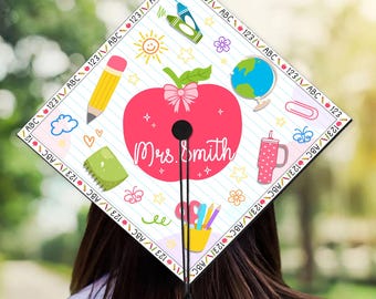 May include: A white graduation cap with a red apple design. The apple has a pink bow and the text "Mrs. Smith" is written on it. The cap is decorated with colorful school-themed doodles, including a pencil, a cloud, a globe, a sun, a flower, a butterfly, a paperclip, a cup, and a ruler.