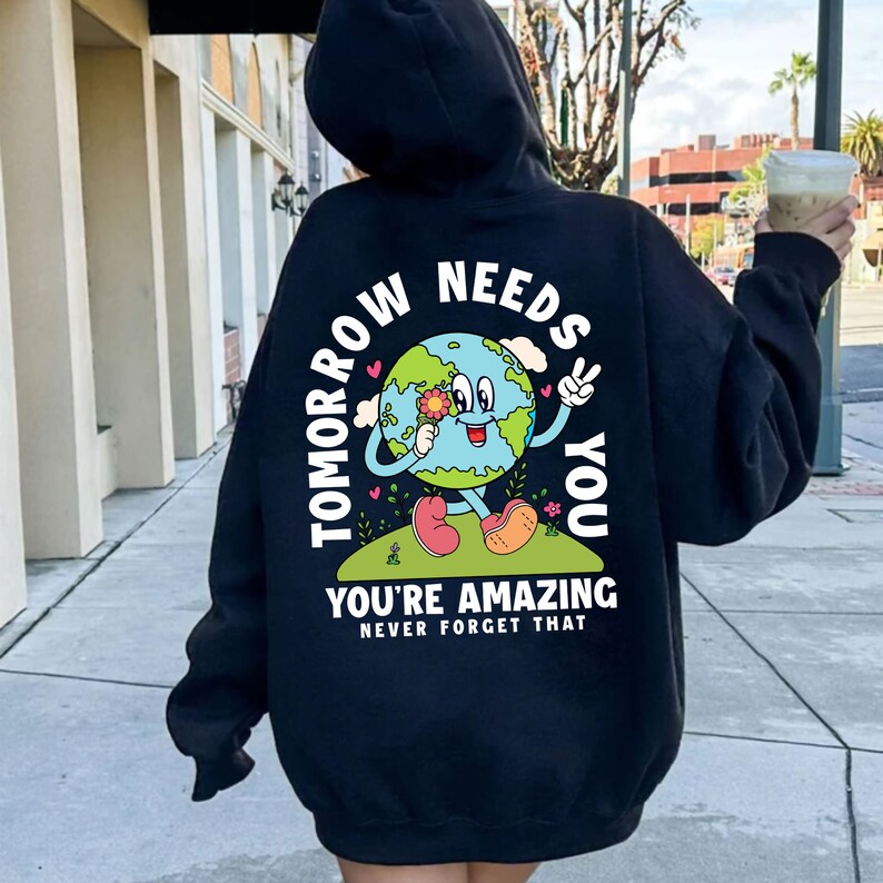 May include: Black hooded sweatshirt with a cartoon Earth graphic wearing a flower and making a peace sign. The text "Tomorrow Needs You" is written around the Earth graphic. The text "You're Amazing Never Forget That" is written below the graphic.