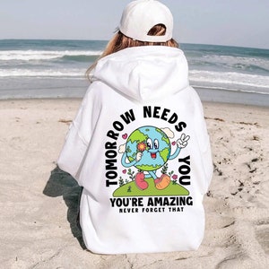 May include: White hoodie with a graphic of a smiling earth holding a flower and the text "Tomorrow Needs You" and "You're Amazing Never Forget That".