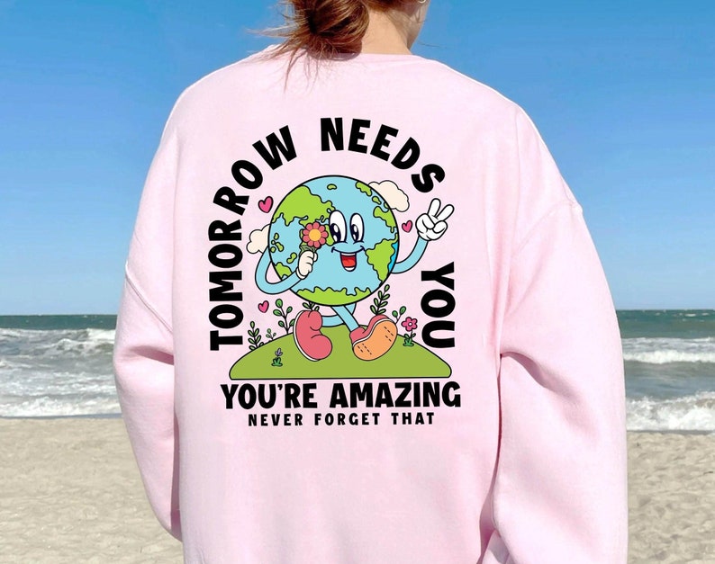 May include: Pink sweatshirt with a graphic of a smiling Earth holding a flower. The text reads "Tomorrow Needs You You're Amazing Never Forget That."