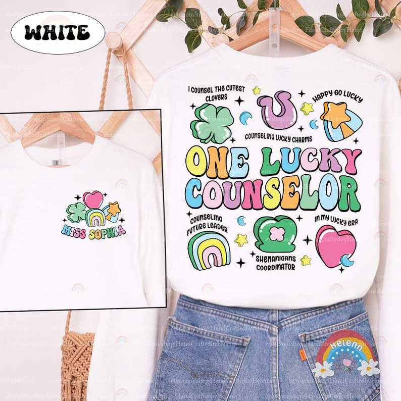 Puede incluir: Camiseta blanca de manga larga con un dise&ntilde;o gr&aacute;fico colorido que incluye el texto "One Lucky Counselor" y varios amuletos de la suerte, como tr&eacute;boles, arco&iacute;ris, estrellas y corazones. El texto "Miss Sophia" est&aacute; en el lado izquierdo de la camiseta.