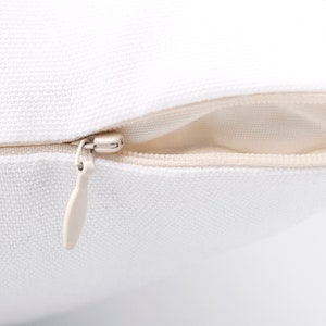 May include: Close-up of a white pillow with a beige zipper. The pillow has a textured surface, and the zipper pull is a matching beige color. The zipper is partially open, revealing the pillow's interior.