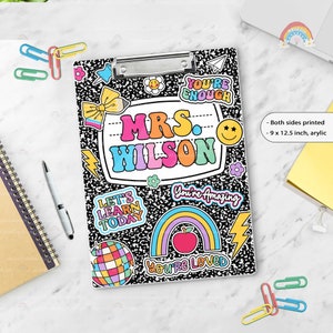Puede incluir: Un portapapeles de acr&iacute;lico de 22,86 x 31,75 cm con un patr&oacute;n de cuaderno de composici&oacute;n en blanco y negro. El centro presenta una etiqueta blanca con el texto MRS. WILSON en letras coloridas, rodeado de pegatinas de arco&iacute;ris, manzana y bola disco.