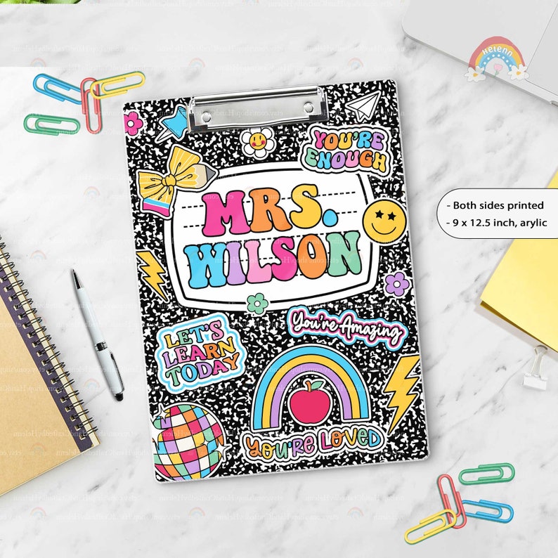 Puede incluir: Un portapapeles de acr&iacute;lico de 22,86 x 31,75 cm con un patr&oacute;n de cuaderno de composici&oacute;n en blanco y negro. El centro presenta una etiqueta blanca con el texto MRS. WILSON en letras coloridas, rodeado de pegatinas de arco&iacute;ris, manzana y bola disco.