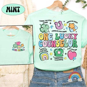 Puede incluir: Una camiseta verde claro con un dise&ntilde;o gr&aacute;fico colorido que dice "One Lucky Counselor". El dise&ntilde;o incluye tr&eacute;boles, arco&iacute;ris, corazones y estrellas. El texto "Miss Sophia" est&aacute; en la parte delantera de la camiseta.