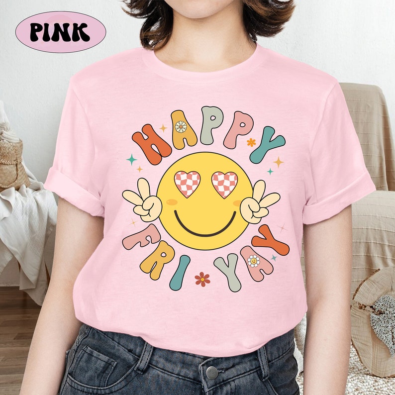May include: Pink t-shirt with a retro-style graphic design featuring a smiley face with checkered heart eyes and the text "Happy Fri Yay" in a colorful, rounded font.