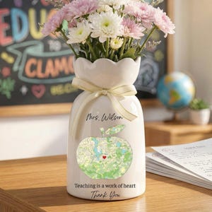 May include: A white ceramic vase with a scalloped top, tied with a cream ribbon, and filled with pink and white flowers. The vase features an apple map design and the text "Mrs. Wilson", "Teaching is a work of heart", and "Thank You".