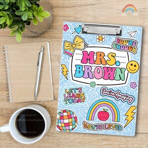Puede incluir: Un portapapeles azul claro con un patr&oacute;n de motas blancas y pegatinas coloridas, incluyendo un arco&iacute;ris, una manzana, una bola de discoteca y el texto Mrs. Brown, You're Enough, Let's Learn Today, You're Amazing y You're Loved.