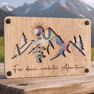 Könnte beinhalten: Eine rechteckige Holztafel mit einem lasergeschnittenen Bergmassiv, Tannenbäumen und einem Wegweiser. Der Ausschnitt gibt den Blick auf einen 20-Euro-Schein hinter dem Holz frei. Der Text "Für dein nächstes Abenteuer" ist unten eingraviert.