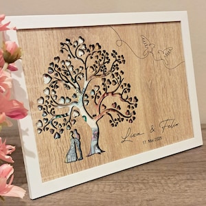 Könnte beinhalten: Eine weiße gerahmte Holzplakette mit einem ausgeschnittenen Baumdesign. Der Baum ist mit Geld gefüllt und zwei Figuren stehen darunter. Der Text "Lisa & Felix 17. Mai 2025" ist unter dem Baum graviert.