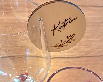May include: A wooden place card with the name "Katrin" engraved on it. The card is attached to a wine glass with a small metal clip. The card has a simple design with a small branch of leaves.