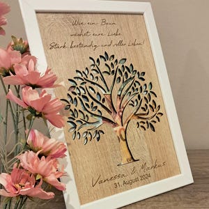 Könnte beinhalten: Eine weiße gerahmte Holzplakette mit einem ausgeschnittenen Baumdesign. Der Baum ist mit Euro-Scheinen gefüllt. Die Plakette trägt den Text "Wie ein Baum wächst eure Liebe Stark, beständig und voller Leben!" und "Vanessa & Markus 31. August 2024".