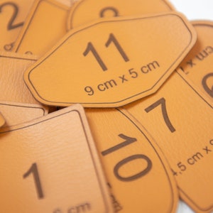 Könnte beinhalten: Nahaufnahme von mehreren braunen Lederetiketten mit Nummern und Maßen. Die Etiketten liegen übereinander, wobei die Nummer "11" und das Maß "9 cm x 5 cm" auf dem obersten Etikett sichtbar sind.