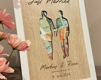 May include: A white wooden frame with a cutout of two people holding hands. The cutout is filled with euro bills. The frame has the text "Just Married", "Markus & Rene", and "8. Juni 2024" engraved on it.