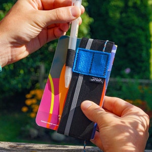 May include: A black pen holder with a blue and gray elastic strap and the word "JAVA" printed on it. The holder is attached to a colorful card with a pen inserted into the holder.