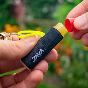 以下が含まれることがあります： 黄色い紐と赤いキャップの小さな円筒形の容器が付いた黒いキーホルダー。容器には「JAVA」と表示され、黄色のリップバームのスティックが入っています。