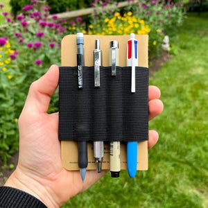 May include: A tan pocket notebook featuring a black elastic pen holder band attached to the cover. The band secures four writing instruments: a black mechanical pencil, a silver metal pen, a white fine-liner pen, and a multi-colour ink pen.