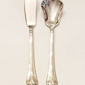 May include: A set of two silver-plated butter knives with ornate handles. The butter knife on the left has a straight blade, while the butter knife on the right has a scalloped edge.