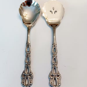 May include: Two silver serving utensils with ornate handles. One is a spoon with a scalloped bowl, and the other is a server with three decorative cutouts. The handles feature a detailed, openwork design.