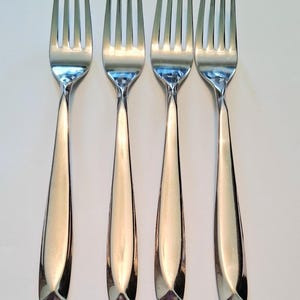 May include: A set of four identical stainless steel dinner forks arranged in a vertical row. Each fork features a polished, reflective finish, a four-tined head, and a smooth, tapered handle that widens slightly toward the base with a flat end.