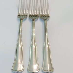 May include: Three silver forks with a polished finish. Each fork has four tines and a handle that widens towards the base. The forks are arranged in a row against a white background, showcasing their reflective surfaces.