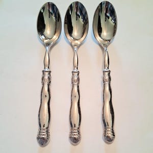 May include: Three silver spoons with ornate handles. The spoons have a classic design, with a rounded bowl and a detailed, decorative handle. The handles have a bulbous shape with intricate patterns. The spoons are arranged on a white surface.