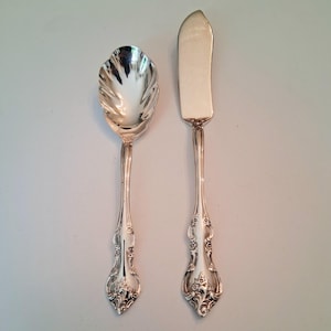 May include: A silver-toned metal serving spoon with a scalloped bowl and a silver-toned metal butter knife. Both utensils feature ornate, embossed floral detailing on the handles, with a polished, reflective finish throughout the entire surface.