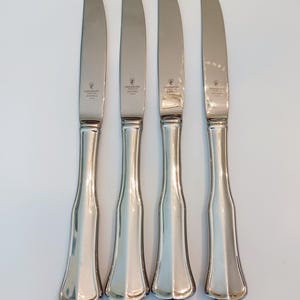 May include: Four stainless steel butter knives with curved blades and ornate handles. Each knife has a silver finish and a decorative design on the handle. The blades are smooth and shiny, reflecting the light. The knives are arranged vertically.