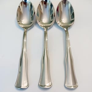 May include: Three silver spoons with a reflective, polished surface. The spoons have a classic design with elongated handles and rounded bowls. The spoons are arranged on a white surface, reflecting the light.