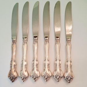 May include: A set of six silver-toned dinner knives arranged vertically. Each knife features a smooth, polished blade and a decorative handle with intricate embossed floral and scrollwork patterns extending from the bolster to the base.