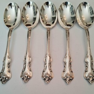 May include: A set of five polished silver-tone spoons arranged in a row. Each spoon features a reflective oval bowl and a decorative handle with intricate embossed floral and scrollwork patterns extending from the base to the neck of the spoon.