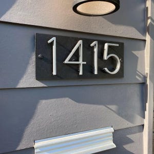 May include: A black rectangular address plaque displaying the number 1415 in silver, mounted on a grey exterior wall. The numbers are in a modern font and are raised from the plaque's surface. A light fixture is above the plaque.