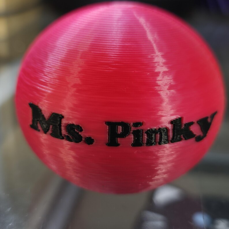 Op de afbeelding: Een helderrode bol met de tekst "Ms. Pinky" in zwart. De bol heeft een getextureerd oppervlak en de tekst is duidelijk gedrukt. Het object lijkt een decoratief item of een onderdeel van een groter product te zijn.