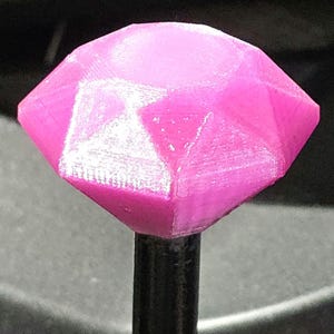 Op de afbeelding: Een roze, gefacetteerd, diamantvormig object op een zwarte staaf. De diamant heeft meerdere platte oppervlakken die het licht vangen. Het object lijkt 3D-geprint, met zichtbare laaglijnen. Het ontwerp is geometrisch en modern.