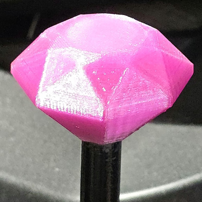 Op de afbeelding: Een roze, gefacetteerd, diamantvormig object op een zwarte staaf. De diamant heeft meerdere platte oppervlakken die het licht vangen. Het object lijkt 3D-geprint, met zichtbare laaglijnen. Het ontwerp is geometrisch en modern.