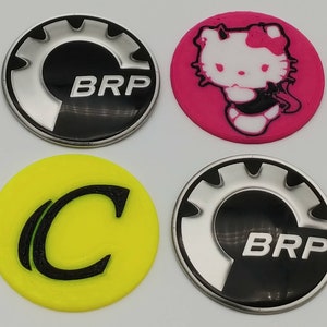 K&ouml;nnte beinhalten: Vier runde Aufkleber mit verschiedenen Designs. Der erste Aufkleber ist schwarz mit einem silbernen Zahnrad und dem Text "BRP". Der zweite Aufkleber ist pink mit einer wei&szlig;en Comicfigur mit dem Text "Hello Kitty". Der dritte Aufkleber ist gelb mit einem schwarzen Buchstaben "C". Der vierte Aufkleber ist schwarz mit einem silbernen Zahnrad und dem Text "BRP".