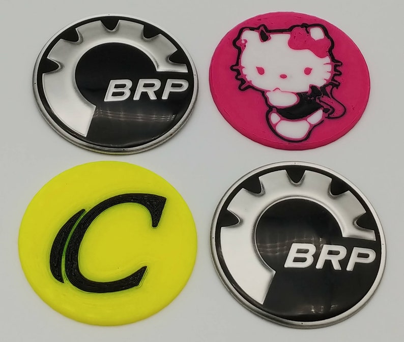 K&ouml;nnte beinhalten: Vier runde Aufkleber mit verschiedenen Designs. Der erste Aufkleber ist schwarz mit einem silbernen Zahnrad und dem Text "BRP". Der zweite Aufkleber ist pink mit einer wei&szlig;en Comicfigur mit dem Text "Hello Kitty". Der dritte Aufkleber ist gelb mit einem schwarzen Buchstaben "C". Der vierte Aufkleber ist schwarz mit einem silbernen Zahnrad und dem Text "BRP".