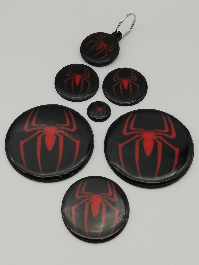 K&ouml;nnte beinhalten: Satz mit sechs runden Schl&uuml;sselanh&auml;ngern mit Spider-Man-Logo in Schwarz und Rot. Die Schl&uuml;sselanh&auml;nger haben eine gl&auml;nzende Oberfl&auml;che. Das Spider-Man-Logo ist rot und befindet sich mittig auf jedem Schl&uuml;sselanh&auml;nger. Die Schl&uuml;sselanh&auml;nger sind in verschiedenen Gr&ouml;&szlig;en.