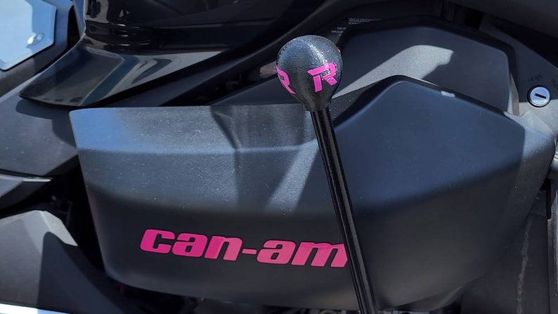 Op de afbeelding: Zwart voertuigonderdeel met de roze tekst "can-am". Een zwarte versnellingspook met een roze "R" logo is zichtbaar. De afbeelding toont mechanische onderdelen van een voertuig.