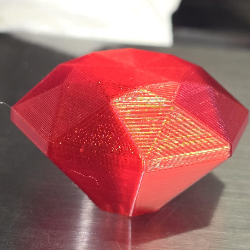 Op de afbeelding: Een gefacetteerde, rode edelsteen met een diamantachtige vorm. De edelsteen heeft een glanzende afwerking en lijkt van een massief materiaal te zijn gemaakt. Het oppervlak is verdeeld in meerdere vlakke vlakken, die licht reflecteren.