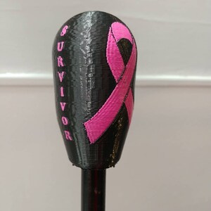 Op de afbeelding: Een zwart, 3D-geprint object met een roze lint en het woord "SURVIVOR" in roze. Het object is gemonteerd op een zwarte staaf. Het roze lint is een symbool van borstkankerbewustzijn.