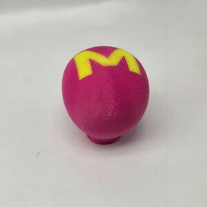 Op de afbeelding: Een magenta-kleurige, bolvormige knop met een geel "M"-ontwerp aan de bovenkant. De knop heeft een afgeronde basis en is waarschijnlijk een decoratief of functioneel element voor meubels of een vergelijkbaar item.
