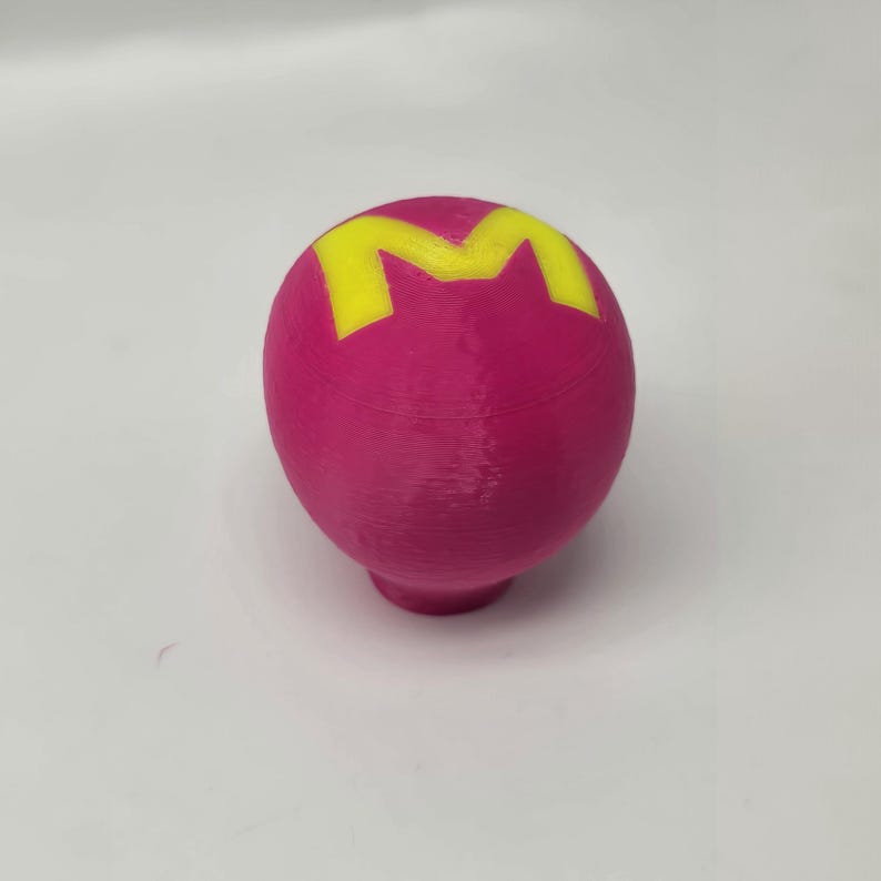 Op de afbeelding: Een magenta-kleurige, bolvormige knop met een geel "M"-ontwerp aan de bovenkant. De knop heeft een afgeronde basis en is waarschijnlijk een decoratief of functioneel element voor meubels of een vergelijkbaar item.