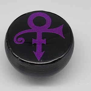 Op de afbeelding: Een zwart, rond object met een paars symbool. Het symbool is een combinatie van mannelijke en vrouwelijke symbolen, met een pijl naar beneden. Het object heeft een glanzende afwerking en is waarschijnlijk decoratief.