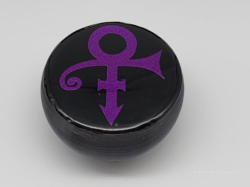 Op de afbeelding: Een zwart, rond object met een paars symbool. Het symbool is een combinatie van mannelijke en vrouwelijke symbolen, met een pijl naar beneden. Het object heeft een glanzende afwerking en is waarschijnlijk decoratief.